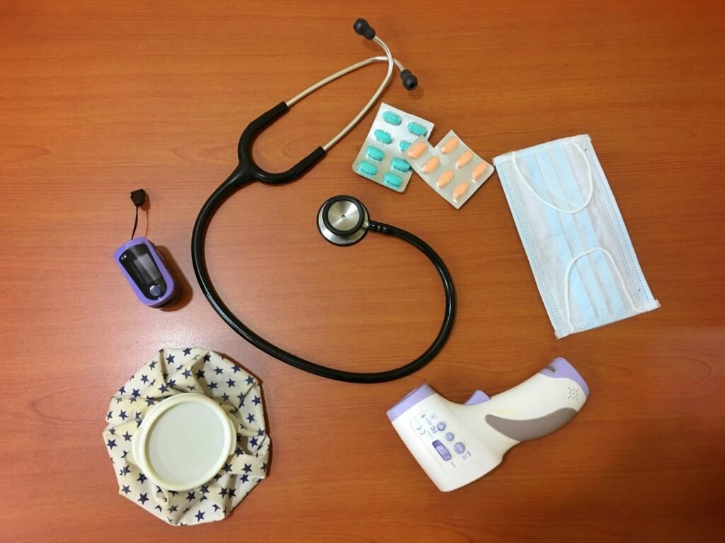 Medical tools on a wooden surface including a stethoscope, digital thermometer, pulse oximeter, face mask, pills, and ice pack.