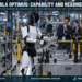Tesla Optimus-style humanoid robot handling an automotive component in a controlled factory while engineers evaluate reliability, safety and commercial readiness.